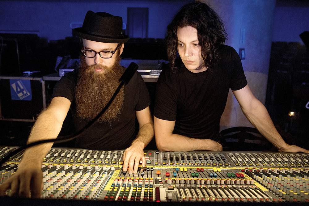 Jack White (right, with long dark hair) and his audio engineer, Joshua V. Smith (left, wearing a black fedora and large beard), lean over a large, multi-channel analog mixing console in a dimly lit studio. Both are wearing black shirts and are focused intently on the controls