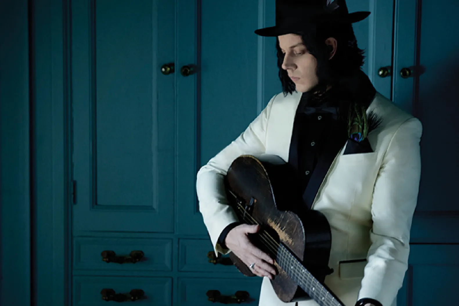 Jack White in a striking pose, wearing a white suit jacket with a black lapel, a black fedora, and a peacock feather accent. He is holding a heavily worn, dark vintage acoustic guitar across his body in a dimly lit, moody blue room, emphasizing the rustic and analog nature of his music.